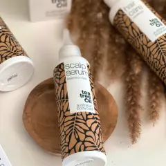 Flera flaskor med "tea tree oil" hårvårdsprodukter, inklusive en "scalp serum" treatment mask, arrangerade med torkade grässtrån.