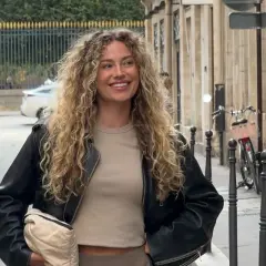 Kvinna med fantastiskt lockigt hår, klädd i skinnjacka och beige topp, ler glatt utomhus.