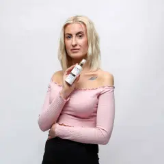 Blond kvinna håller upp en flaska med hårvårdsprodukt, kanske ett silikon schampo, framför en vit bakgrund.