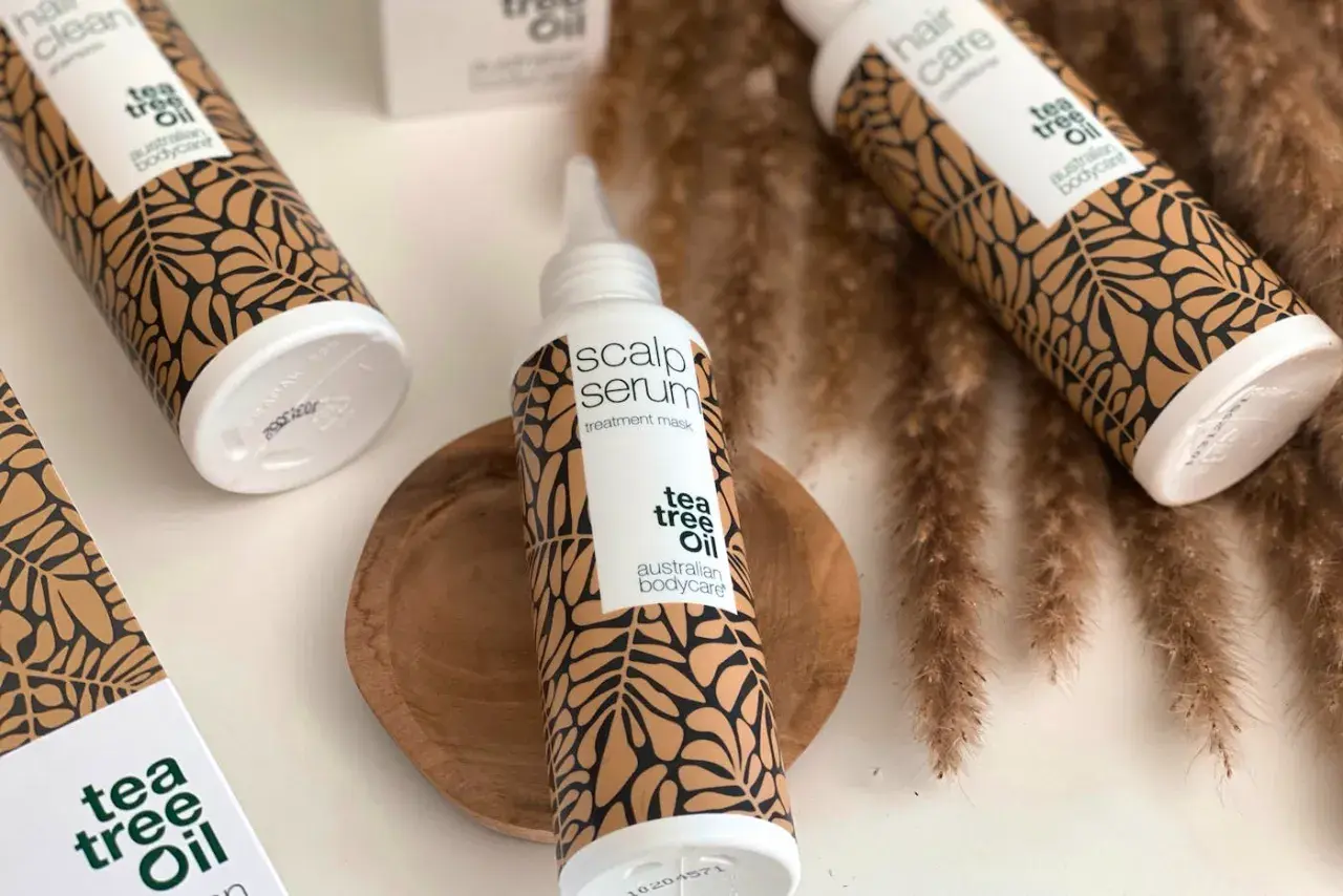 Flera flaskor med "tea tree oil" hårvårdsprodukter, inklusive en "scalp serum" treatment mask, arrangerade med torkade grässtrån.