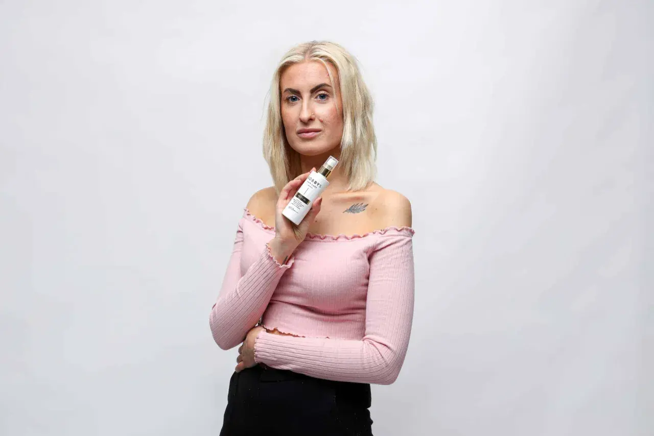 Blond kvinna håller upp en flaska med hårvårdsprodukt, kanske ett silikon schampo, framför en vit bakgrund.